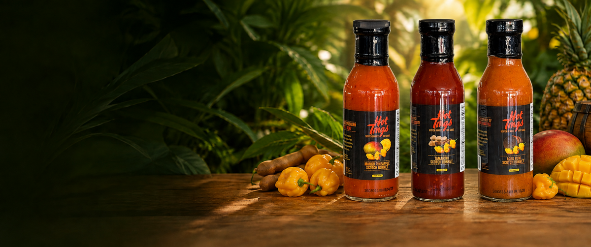 Three bottles of Hot Tings Caribbean Scotch Bonnet hot sauce lined up on a wooden table with fresh tropical fruits and vegetables, set against a lush green leafy background.